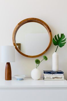 a mirror and some books on a white shelf next to a table with a lamp