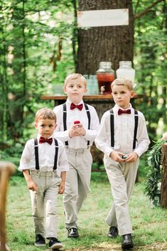 three young boys dressed in white and black bow ties walking through the woods with their hands together