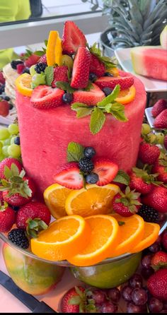 there is a cake with fruit on it and other fruits around the cake, including oranges, strawberries, raspberries, lemons, and watermelon