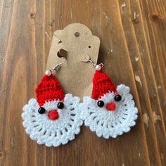 two red and white crocheted santa claus earrings on a wooden table next to a tag