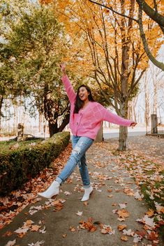 a woman in pink jacket and jeans dancing on sidewalk surrounded by trees with leaves around her