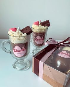 two mugs filled with hot chocolate and whipped cream, next to a gift box