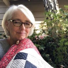 an older woman with glasses is holding a quilt in front of a tree and bushes