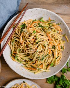 a white bowl filled with noodles and veggies next to chopsticks on a wooden table