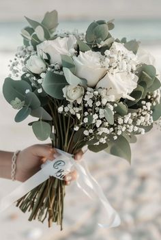 a woman holding a bouquet of white flowers and greenery in her hand on the beach