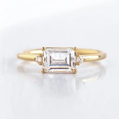 a gold ring with an emerald cut diamond and three diamonds on the band, sitting on a white surface