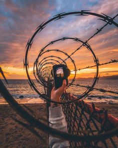 a person taking a photo with their cell phone through barbed wire on the beach at sunset