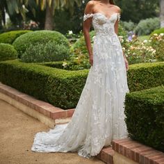 a woman in a white wedding dress standing next to some bushes and trees with her hands on her hips