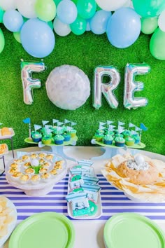 a table topped with lots of food next to green plates and blue and white balloons