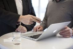 two men sitting at a table with an apple laptop in front of them, one pointing to the screen