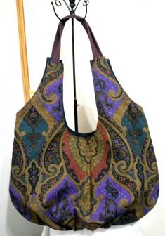 Large hobo bag with boho colors and features is 13.5" high at center of bag above the outside zipper by 21" wide at its widest point. The bag is made of very smooth decorator weight fabric.  The colors are subdued black, tan, purple and plum. There are 2 zipper pockets; one exterior and one interior.  The plum colored handle fits the purse perfectly.   Bag is made from the Diana Tote Bag Pattern by Delinda Boutique. Lining is a shiny denim. Use for book bag, knitting or crochet bag, lunch bag, gift bag, shopping bag, everyday bag or whatever you need it for. Bohemian Reversible Shoulder Bag, Bohemian Reversible Hobo Bag For Daily Use, Bohemian Multicolor Reversible Bag, Bohemian Reversible Hobo Tote Bag, Bohemian Reversible Hobo Bag For Everyday Use, Multicolor Rectangular Bag With Paisley Print, Bohemian Reversible Hobo Bag For Travel, Reversible Bohemian Shoulder Bag, Bohemian Multicolor Tapestry Bags