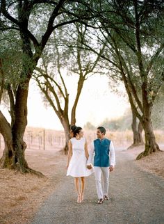 a man and woman holding hands walking down a dirt road in front of some trees