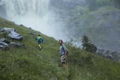 two people are hiking up a hill near a waterfall in the distance, with trees and grass on either side