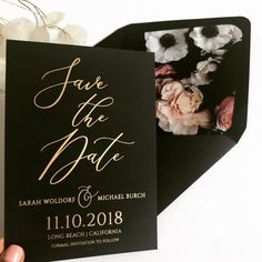 a black and gold save the date card with white flowers on it is held up by someone's hand