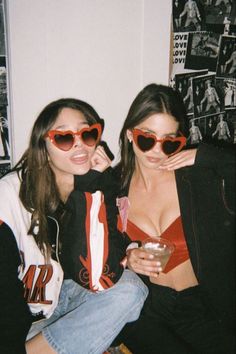 two women wearing heart shaped sunglasses sitting next to each other in front of a wall