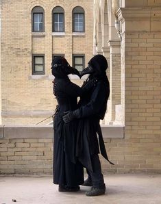 two people dressed in black standing next to each other near a brick building with arched windows