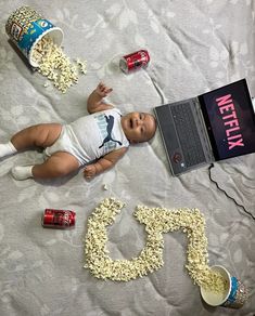 a baby laying on top of a bed next to a laptop computer and some popcorn