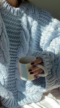 a woman is holding a coffee cup in her hand while sitting on a bed wearing a sweater