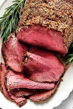 sliced roast beef on a white plate with rosemary sprigs and seasoning next to it