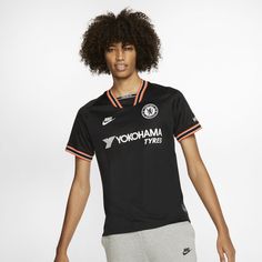 a young man with curly hair wearing a black shirt and grey sweatpants, standing in front of a white background