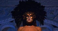 a woman with black feathers on her head wearing a brown dress and gold jewelry in front of a blue wall