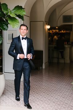 a man in a tuxedo and bow tie is walking towards the camera with his hands in his pockets