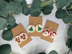 three wooden earrings with christmas designs on them sitting next to some green leafy plants