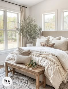 a large bed sitting in a bedroom next to two windows and a table with pillows on it