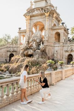 two women standing next to each other in front of a building with a fountain on it