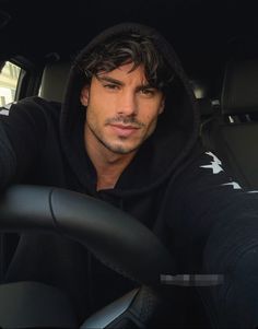 a man sitting in the drivers seat of a car, wearing a hoodie and looking at the camera