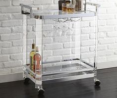 a clear bar cart with wine glasses and bottles on the bottom shelf in front of a brick wall