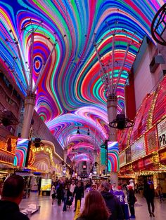 people are walking through an indoor shopping mall with brightly colored lights on the walls and ceiling