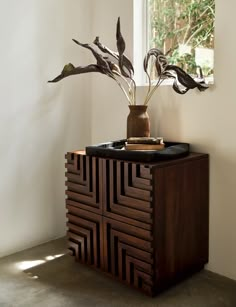 a vase with some plants in it on top of a wooden cabinet next to a window