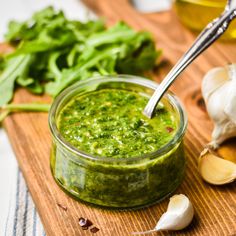 a wooden cutting board topped with a jar of pesto sauce next to garlic and herbs