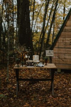 a wooden table topped with a cake next to a small cabin in the middle of a forest