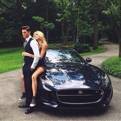 a man and woman leaning on the hood of a sports car in front of trees