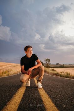 a man squatting down on the side of an empty road with clouds in the background