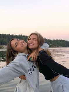 two young women standing next to each other in front of the water with their arms around one another