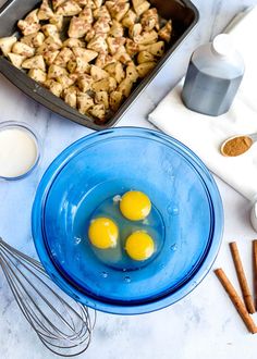 three eggs in a blue bowl next to cinnamon sticks and other ingredients on a white counter