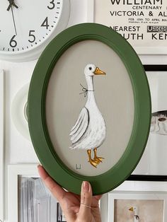 a hand holding up a framed painting of a duck in front of a wall clock