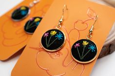 two pairs of earrings with flowers on them sitting next to an orange piece of paper