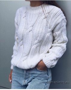 a woman wearing a white sweater and jeans with her hands in her pockets, leaning against a wall