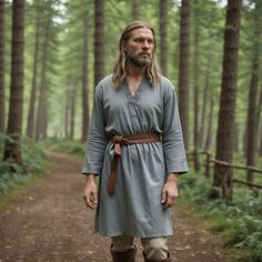 a man with long hair and beard standing in the middle of a forest wearing a gray dress