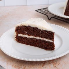 a slice of chocolate cake with white frosting sits on a plate next to the rest of the cake