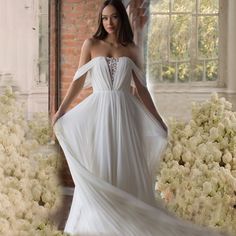 a woman in a white dress standing next to flowers and brick wall with large windows