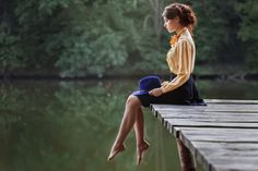 a woman sitting on a dock with her legs crossed looking at the water and trees in the background