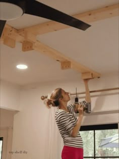 a woman in striped shirt and red pants working on a ceiling fan with black blades