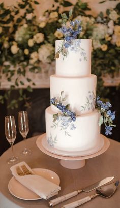 a three tiered wedding cake with blue flowers on the top and two wine glasses next to it