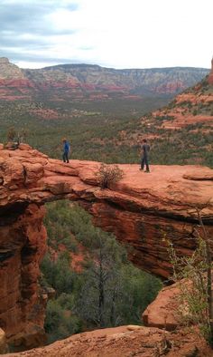 two people are standing on the edge of a cliff looking out at trees and mountains