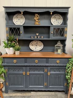 a blue china cabinet with plates on top and plants in the corner next to it